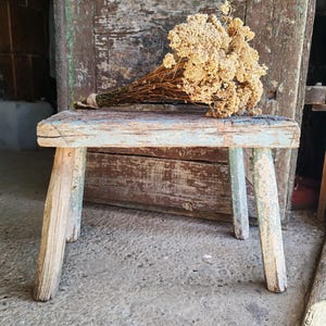 Puede incluir: Un taburete de madera pequeño y desgastado con cuatro patas y un asiento rectangular que presenta restos de pintura azul claro. Sobre el asiento descansa un ramo seco de pequeñas flores de color beige atadas con un trozo de cuerda natural.