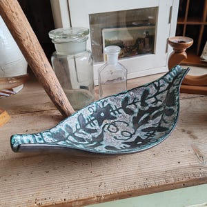 May include: A leaf-shaped ceramic dish featuring a textured surface with a dark black and teal green glaze. The interior displays a stylized, etched botanical pattern with intricate leaf and vine motifs across the curved, elongated form.