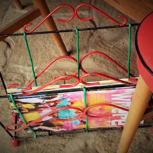 May include: A retro magazine rack with a black metal frame and red and green detailing. The rack is filled with magazines with vibrant covers. A red chair is partly visible in the background.