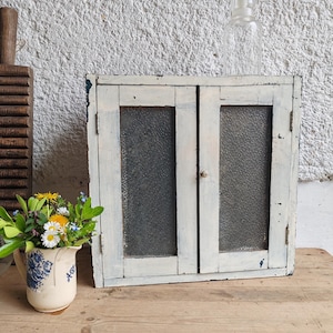 May include: A small, vintage white cabinet with two glass-paned doors. The cabinet has a distressed, weathered appearance, with visible wood grain and peeling paint. A clear glass bottle sits on top. A floral pitcher is on the left.