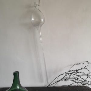 May include: A clear glass bulb with a long, thin neck hanging from a white wall. The bulb is empty and has a rounded shape. The neck of the bulb is very long and thin, and it is hanging straight down.