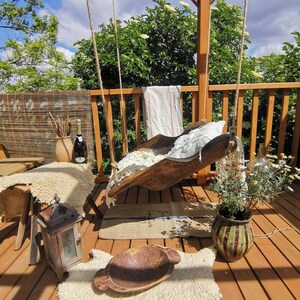 May include: A wooden swing seat hangs from a wooden pergola. The swing seat is made of a natural wood and has a white fabric cushion. The swing seat is hanging over a wooden deck with a brown rug and a potted plant.