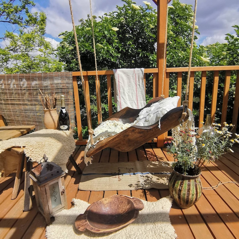 May include: A wooden swing seat hangs from a wooden pergola. The swing seat is made of a natural wood and has a white fabric cushion. The swing seat is hanging over a wooden deck with a brown rug and a potted plant.