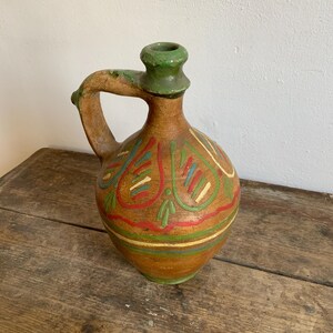 May include: A brown ceramic pitcher with a green top and a handle. The pitcher is decorated with colorful geometric patterns.