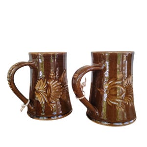 May include: Two brown ceramic mugs with handles. Each mug features a raised floral design and vertical stripes. The handles are attached with a natural fibre string. The mugs are glazed with a glossy finish.