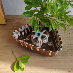 May include: A brown ceramic flower frog with a light blue interior and a brown and white exterior. The frog is placed inside a brown ceramic dish with a light blue interior and brown and white exterior. Green leaves and branches are placed in the frog.