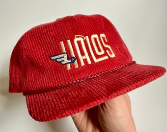 Puede incluir: Una gorra de béisbol de pana roja con un halo y alas bordadas en oro sobre la palabra "HALOS" en bordado dorado.