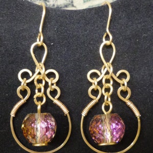 May include: A pair of gold-toned wire earrings featuring a dangling faceted purple bead suspended within a circular wire frame. The upper portion of each earring consists of intricate wire loops and coils, finished with a standard hook ear wire.