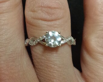 May include: A silver-toned ring featuring a round, light blue center stone held by four prongs. The band has a twisted design with small, clear, sparkling stones embedded along the metal surface on both sides of the central gemstone.