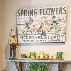 Może przedstawiać: Rusztykalny drewniany znak z tekstem "SPRING FLOWERS" i "PICKED FRESH DAILY" z napisami "PEONIES ROSES LILACS" poniżej. Znak przedstawia wz&oacute;r kwiatowy z r&oacute;żowymi r&oacute;żami i zielonymi liśćmi. Ceramiczna figurka kr&oacute;lika trzymająca małą łopatę znajduje się na p&oacute;łce pod znakiem z kilkoma szklanymi świecznikami.
