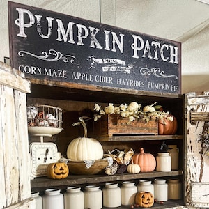 Könnte beinhalten: Ein rechteckiges Schild mit dem Text PUMPKIN PATCH, CORN MAZE, APPLE CIDER, HAYRIDES, PUMPKIN PIE und PICK YOUR OWN. Darunter befindet sich ein Holzschrank mit einem weißen Kürbis, orangefarbenen Kürbissen, zwei Kürbislaternen und weißen Keramikgefäßen.