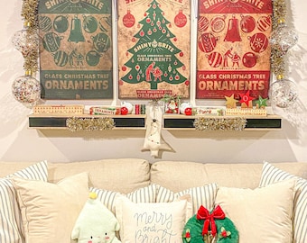 May include: Three rectangular wall signs featuring vintage Shiny Brite glass Christmas tree ornament graphics in green, cream, and red. Below, two shelves hold small boxes and decor. The sofa features cream pillows, a plush tree, a wreath, and a pillow with the text merry and bright.