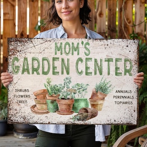 May include: A white sign with green text that reads "Mom's Garden Center". The sign has a distressed, vintage look and features illustrations of potted plants. The sign also lists categories of plants, including shrubs, flowers, trees, annuals, perennials, and topiaries.