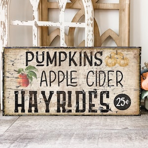 Pode incluir: Uma placa retangular com um fundo creme desgastado apresentando texto em preto que diz PUMPKINS, APPLE CIDER e HAYRIDES. A placa inclui um gráfico de uma maçã vermelha com folhas verdes, três pequenas abóboras amarelas e um círculo preto com 25 centavos.