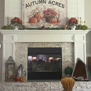 May include: A white fireplace mantel with a stone surround. The mantel is decorated with fall-themed decor, including a sign that reads "Autumn Acres pick your own pumpkins mums apples", a wreath, and a lantern.