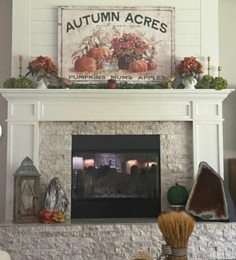 May include: A white fireplace mantel with a stone surround. The mantel is decorated with fall-themed decor, including a sign that reads "Autumn Acres pick your own pumpkins mums apples", a wreath, and a lantern.