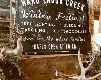 May include: A black rectangular sign with white text reading HARD LABOR CREEK Winter Festival, TREE LIGHTING, SLEDDING, CAROLING, HOT CHOCOLATE, Fun for the whole family!, GATES OPEN AT 10 AM, decorated with a silver deer figurine and antlers.