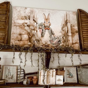 May include: A rectangular canvas print featuring a rabbit wearing a blue jacket and red bowtie, holding carrots and a basket. The print is flanked by two dark brown wooden shutters. Below, a shelf displays two framed prints, a metal rabbit figurine, a small stack of books, and a galvanized metal bucket.
