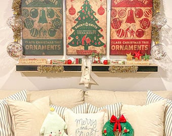 May include: Three vintage-style Shiny Brite glass ornament posters displayed above a beige sofa with decorative pillows. The posters feature red, green, and cream colors with Christmas tree and ornament graphics. Gold tinsel and holly accents frame the wall display.