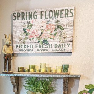 Może przedstawiać: Rustykalny drewniany znak z tekstem "Spring Flowers" i "Picked Fresh Daily" z napisami "Peonies, Roses, Lilacs" poniżej. Znak przedstawia wz&oacute;r kwiatowy z r&oacute;żowymi r&oacute;żami i zielonymi liśćmi. Znak jest umieszczony na p&oacute;łce z ceramiczną figurką kr&oacute;lika i świecami.