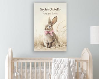 Op de afbeelding: Een rechthoekige wandkunstprint met een bruin konijn met een roze strik om zijn nek, zittend in hoog gras. De tekst "Sophia Isabella" en "you are loved" is in donkerbruin gedrukt boven het konijn.