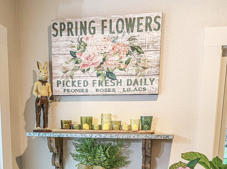 Może przedstawiać: Rusztykalny drewniany znak z tekstem "SPRING FLOWERS" i "PICKED FRESH DAILY" z napisami "PEONIES ROSES LILACS" poniżej. Znak przedstawia wz&oacute;r kwiatowy z r&oacute;żowymi r&oacute;żami i zielonymi liśćmi. Ceramiczna figurka kr&oacute;lika trzymająca małą łopatę znajduje się na p&oacute;łce pod znakiem z kilkoma szklanymi świecznikami.