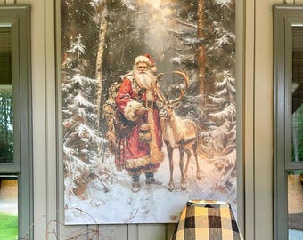 Puede incluir: Un cuadro rectangular que representa a un Santa Claus barbudo con abrigo rojo de pie en un bosque nevado junto a un reno. Santa lleva un saco grande y una linterna, rodeado de pinos cubiertos de nieve bajo una luz suave y brillante.