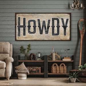 May include: A rectangular wooden wall sign featuring the word HOWDY in large, distressed black capital letters against a weathered, light-coloured wood grain background. The sign is mounted horizontally on a dark grey horizontal plank wall.