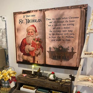May include: A rectangular wall art piece depicting an open book with vintage-style illustrations and text. The left page shows Santa Claus holding a gift, titled A Visit from St. Nicholas. The right page features the poem Twas the night before Christmas.