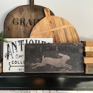May include: A rectangular dark gray sign featuring a light gray silhouette of a running rabbit and the text White Cottage Hare Farm. The sign is positioned in front of a white sign with black text and two wooden pizza peels of different tones.