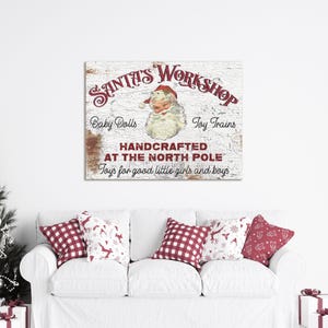 May include: A rectangular wall sign with a distressed white wood texture featuring the red text Santa's Workshop, Baby Dolls, Toy Trains, and Handcrafted at the North Pole. A central illustration shows a vintage Santa Claus face with a red hat.