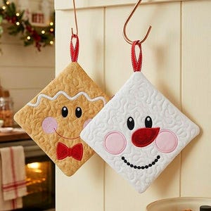 May include: Two diamond-shaped quilted pot holders hanging from copper hooks. One is tan with a gingerbread person face, white icing trim, pink cheeks, and a red bow tie. The other is white with a snowman face, pink cheeks, and a red nose.