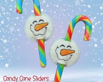 May include: Two candy canes with rainbow stripes and snowman-shaped felt sliders. The snowman has a stitched face and an orange carrot nose. The text "Candy Cane Sliders Set of 6" is at the bottom of the image.