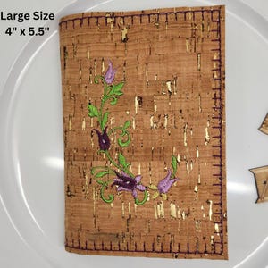 May include: A cork notebook cover with a floral design embroidered in purple, green, and gold. The cover is 4 inches by 5.5 inches.