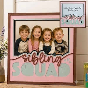 May include: A square wooden photo frame featuring a pink background with the text "sibling SQUAD" in layered script and block lettering. The frame includes a central opening displaying a photograph of four children and a small inset sign.