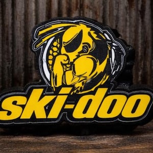May include: A black and yellow Ski-Doo logo, featuring a stylized yellow wasp or hornet design within a white and black border. The word "Ski-doo" is in yellow, outlined in black.