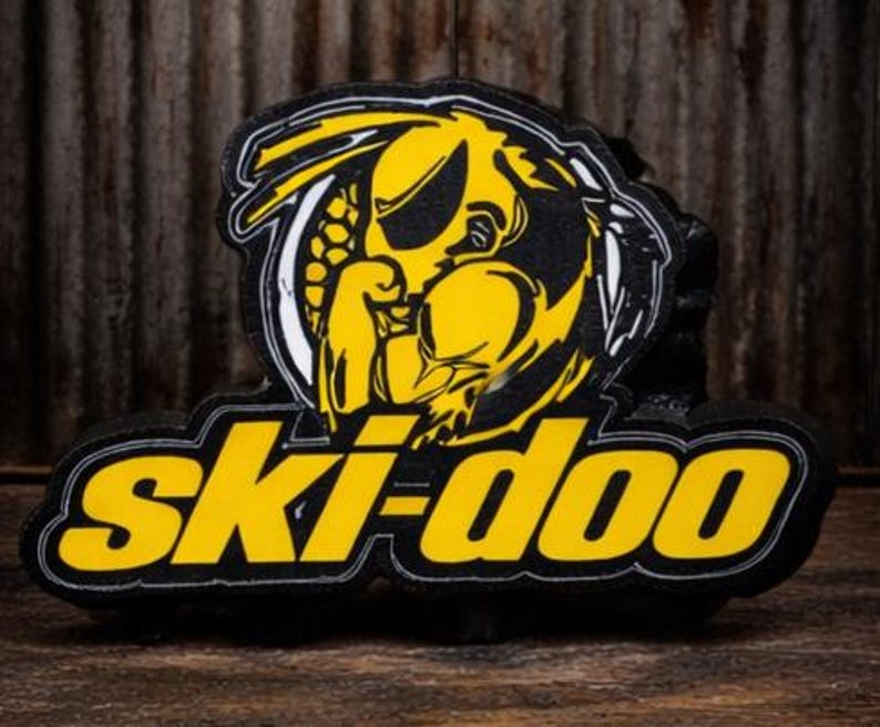 May include: A black and yellow Ski-Doo logo, featuring a stylized yellow wasp or hornet design within a white and black border. The word "Ski-doo" is in yellow, outlined in black.