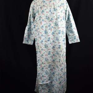 May include: A long-sleeved nightgown in a light blue color featuring a repeating floral pattern of small blue roses and tiny red berries. The garment has a round neckline with a button-down placket at the chest and a straight, loose-fitting silhouette.