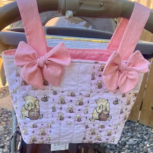 May include: A quilted fabric bag featuring a repeating pattern of Winnie the Pooh characters with honey pots and bees on a light pink background. The bag has two pink straps with white polka dots, each adorned with a large pink bow at the top.