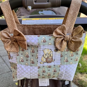 May include: A quilted fabric stroller organizer featuring a Winnie the Pooh pattern with light green, white, and tan colors. The bag has two top handles, each adorned with a tan fabric bow featuring a swirling pattern, and a central patch showing Pooh Bear with a honey pot.