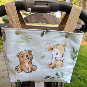 May include: A light blue stroller organizer with a bear and deer design. The organizer has a quilted pattern and is decorated with green leaves. The straps are tan with a swirl pattern. The organizer is attached to a black stroller handle.