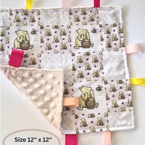May include: A baby security blanket featuring a Winnie the Pooh pattern with a soft, textured corner. The blanket has colorful ribbons and measures 12" x 12".