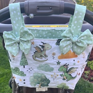 May include: A quilted fabric stroller organizer bag featuring a white background with a green dragon, tree, and cloud print. The bag has two light green polka dot straps with decorative bows attached to the top, hanging from a black stroller handle.