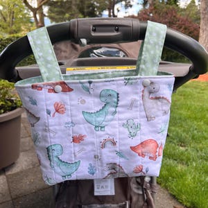 May include: A white and green stroller organizer with a dinosaur print. The organizer has a quilted pattern and is attached to the stroller handle with green straps. The dinosaur illustrations are in shades of blue, red, and gray.