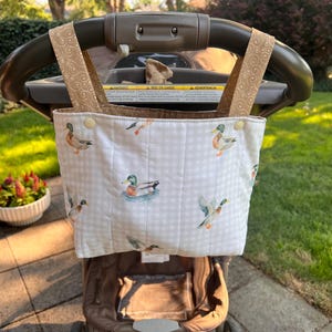 May include: A quilted fabric stroller organizer bag featuring a white and light gray gingham pattern with illustrations of mallard ducks. The bag has two tan straps with a swirling pattern and is attached to a dark gray stroller handle.