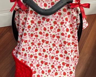 May include: A baby car seat cover featuring a white background with a pattern of red strawberries, pink bows, and flowers. The cover is trimmed with a red, textured blanket and tied with red ribbons. The car seat handle is black.