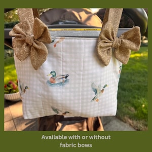 May include: A white quilted fabric pouch featuring a light gray gingham pattern and a repeating mallard duck print. The pouch includes two tan fabric straps with decorative bows featuring a swirling pattern attached to the top corners.