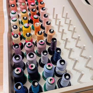 Op de afbeelding: Een witte lade-organizer met meerdere verticale pinnen die verschillende klossen naaigaren vasthouden in een kleurenspectrum van roze, oranje, geel, paars en blauw. De klossen zijn in rijen gerangschikt met enkele lege pinnen zichtbaar.