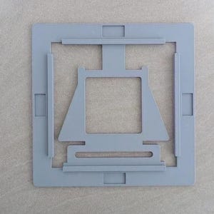 May include: A light grey, square-shaped plastic frame featuring a central, trapezoidal cutout with a vertical support bar extending to the top edge. The frame includes four rectangular indentations along its outer perimeter and raised edges.