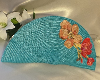 May include: A turquoise, semi-circular clutch with a woven texture. The clutch features an embroidered floral design in shades of orange and red. A silver zipper and white lilies are visible in the background.